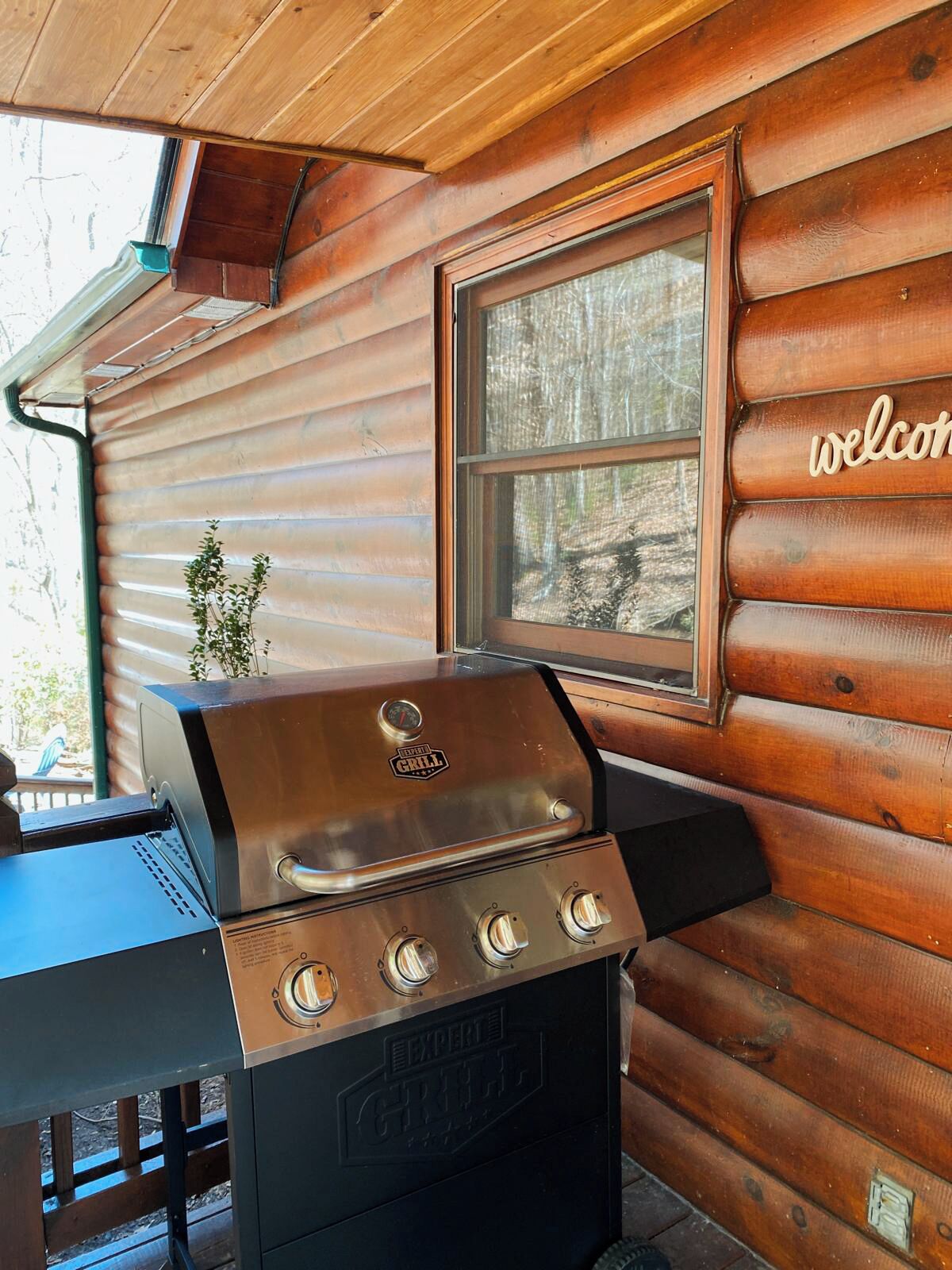 A wooden cabin with a grill and a window with a 'welcome' sign.