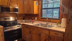 A kitchen with wooden cabinets, a stainless steel stove, a sink, and a window with a view of trees. - Bigfoot's Hideaway | Hot Tub | Fire Pit | Cozy Wooded Retreat (Blue Ridge)