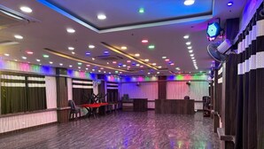 Ein geräumiger Saal mit einem glänzenden Boden, mehreren Deckenleuchten und einer Bühne mit Stühlen und einem Tisch.