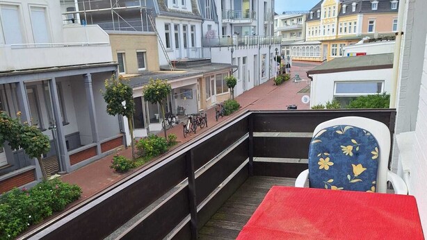 A balcony with a red table and two chairs, overlooking a street with buildings and a flag. - Central vacation apartment Norderney (Norderney)