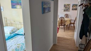 A narrow hallway with a bed on the left, a dining area with chairs and a table in the center, and a chair with a patterned cushion on the right. - Central vacation apartment Norderney (Norderney)