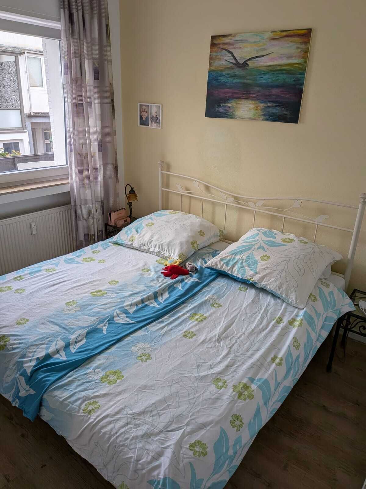 Una cama con ropa de cama floral, un cuadro en la pared y una ventana con cortinas.