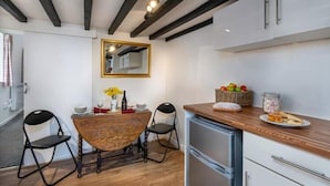 A compact kitchen with a wooden table, two chairs, a refrigerator, and a countertop with food items. - Eisteddfa Country House and Cottages (Criccieth)