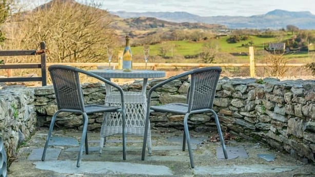 A stone wall with a table and two wicker chairs facing a scenic view of hills and a distant building. - Eisteddfa Country House and Cottages (Criccieth)