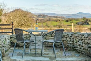 Eine Steinsichtmauer mit einem Tisch und zwei Korbstühlen, die auf eine schöne Aussicht über Hügel und ein entferntes Gebäude blickt.