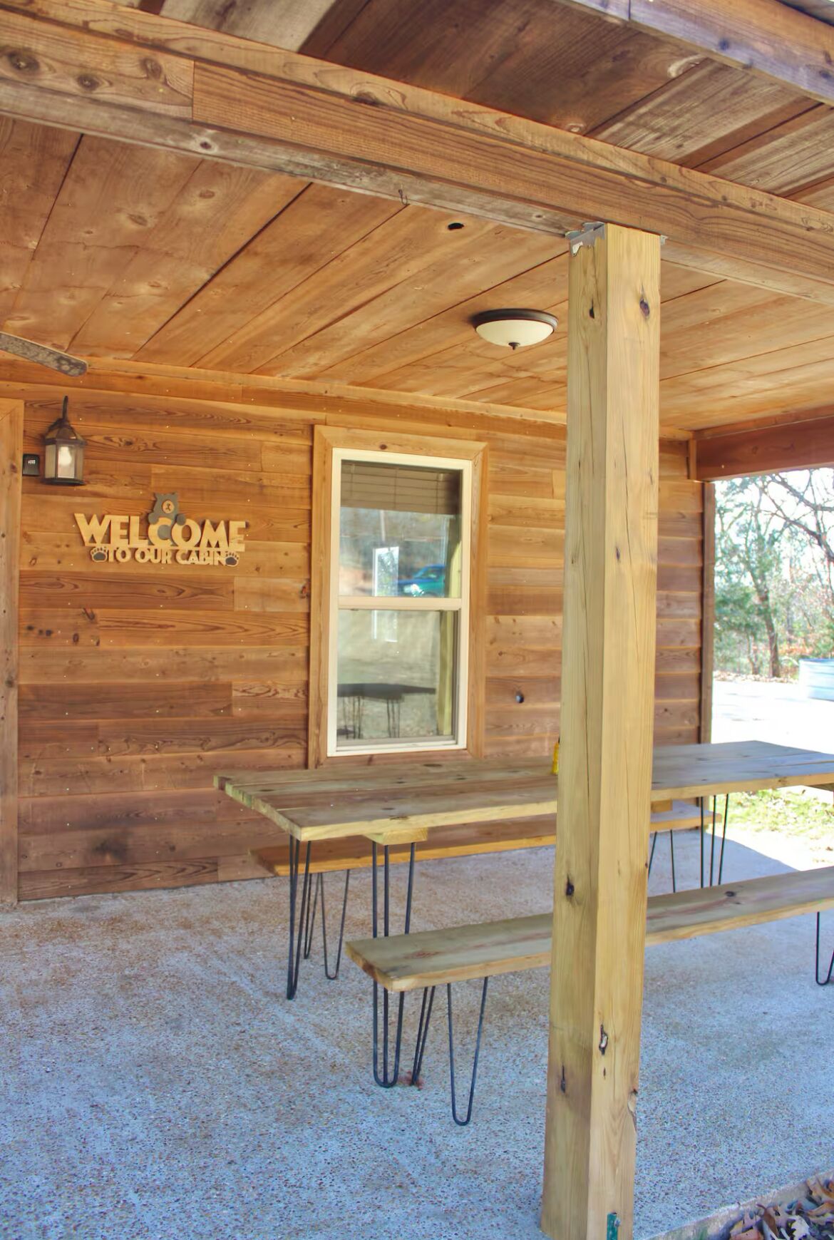 A wooden cabin with a 'WELCOME TO OUR CABIN' sign, a window, and two picnic tables.