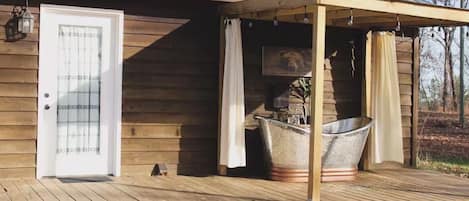Una cabaña de madera con techo metálico, un porche cubierto, una bañera metálica grande y una puerta blanca.