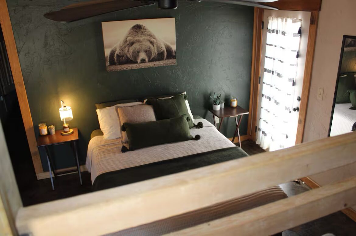 A bedroom with a bed, a nightstand with a lamp, a small table with a plant, and a window with curtains.