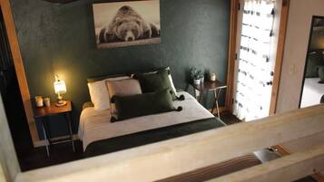 A bedroom with a bed, a nightstand with a lamp, a small table with a plant, and a window with curtains.