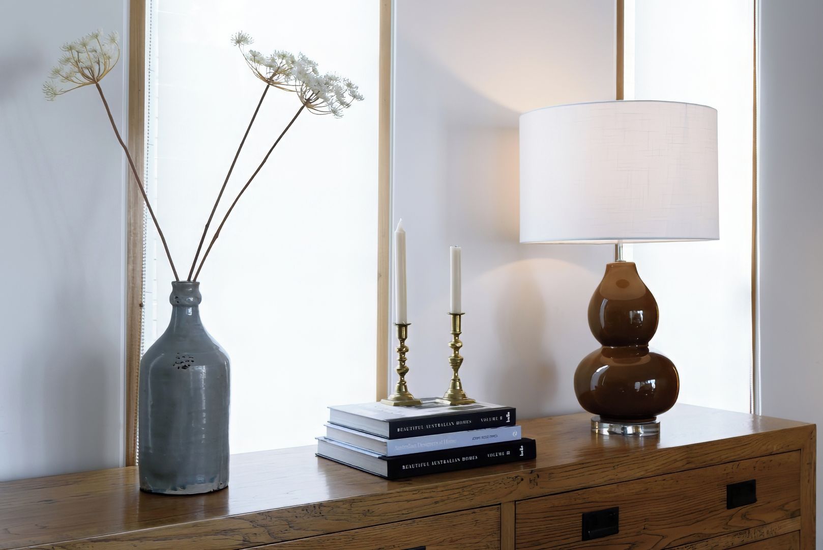 Une commode en bois, sur laquelle on a posé un vase de fleurs séchées, une pile de livres, une bougie et une lampe.