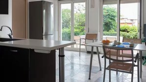 A modern kitchen with a built-in refrigerator, a dining area with a table and chairs, and large sliding glass doors leading to a balcony with a view of greenery. - Terrace Garden Apartment (Aci Trezza)