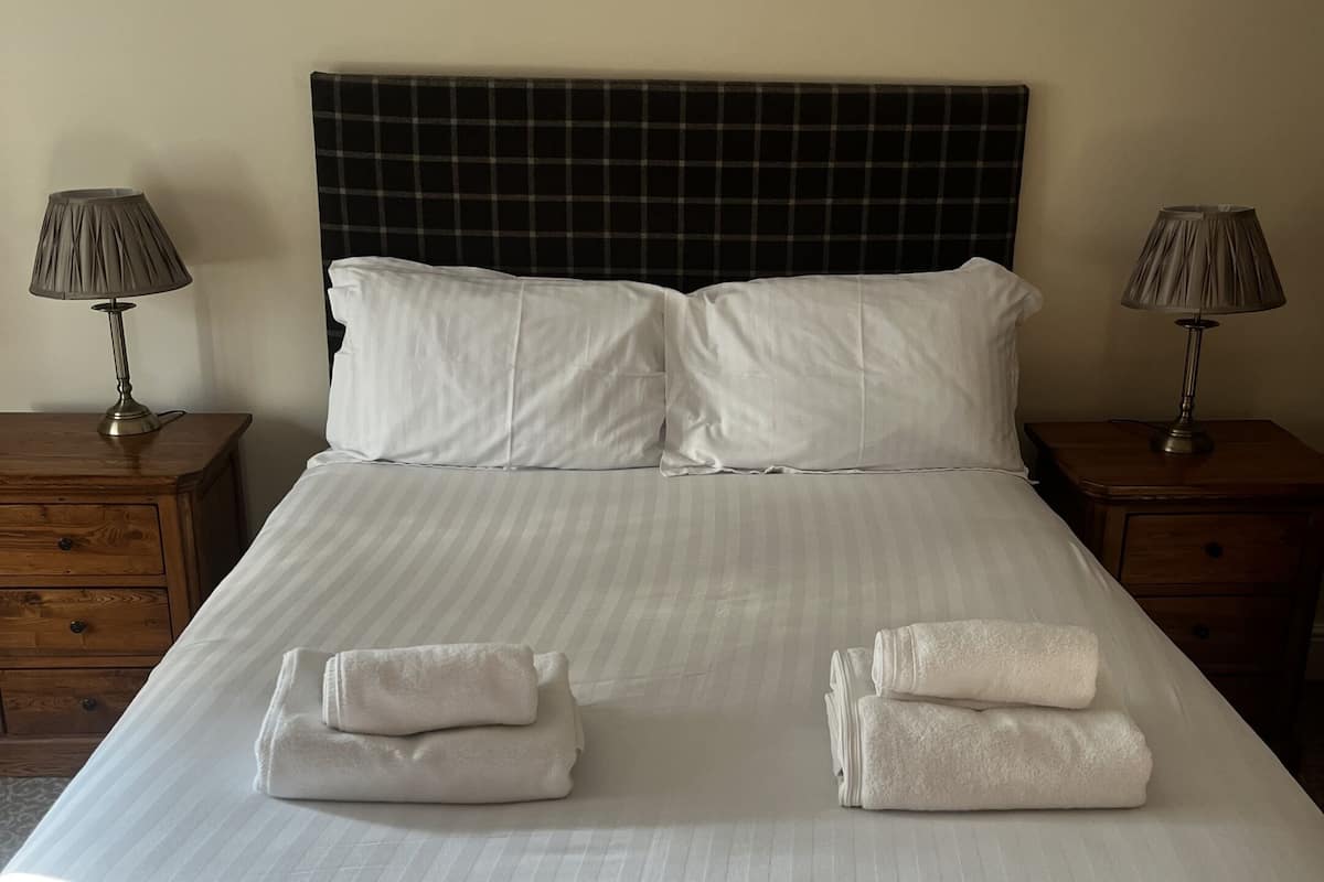 A neatly made bed with white linens and two folded towels on the bed. There are two bedside tables with lamps on either side of the bed.