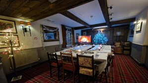 Una sala da pranzo con un lungo tavolo apparecchiato per un pasto, travi in legno al soffitto, un tappeto a quadri e un albero di Natale illuminato.
