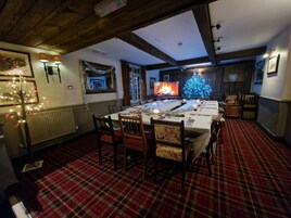 A dining room with a long table set for a meal, wooden beams on the ceiling, a plaid carpet, and a Christmas tree with lights.
