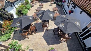 Una terrazza esterna con diversi tavoli e sedie, alcune coperte da ombrelloni, e un edificio bianco con tetto a tegole sullo sfondo.