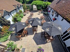 An outdoor patio with multiple tables and chairs, some covered by umbrellas, and a white building with a tiled roof in the background.