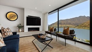A modern living room with a fireplace, a large TV, and a view of a lake and mountains. - The Bunkers Penthouse-2 Bed (Queenstown)