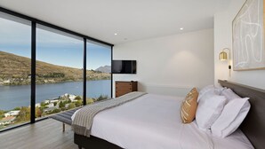 A bedroom with a large window offering a view of a lake and hills, a bed with white linens, a wooden bench, and a mounted TV. - The Bunkers Penthouse-2 Bed (Queenstown)