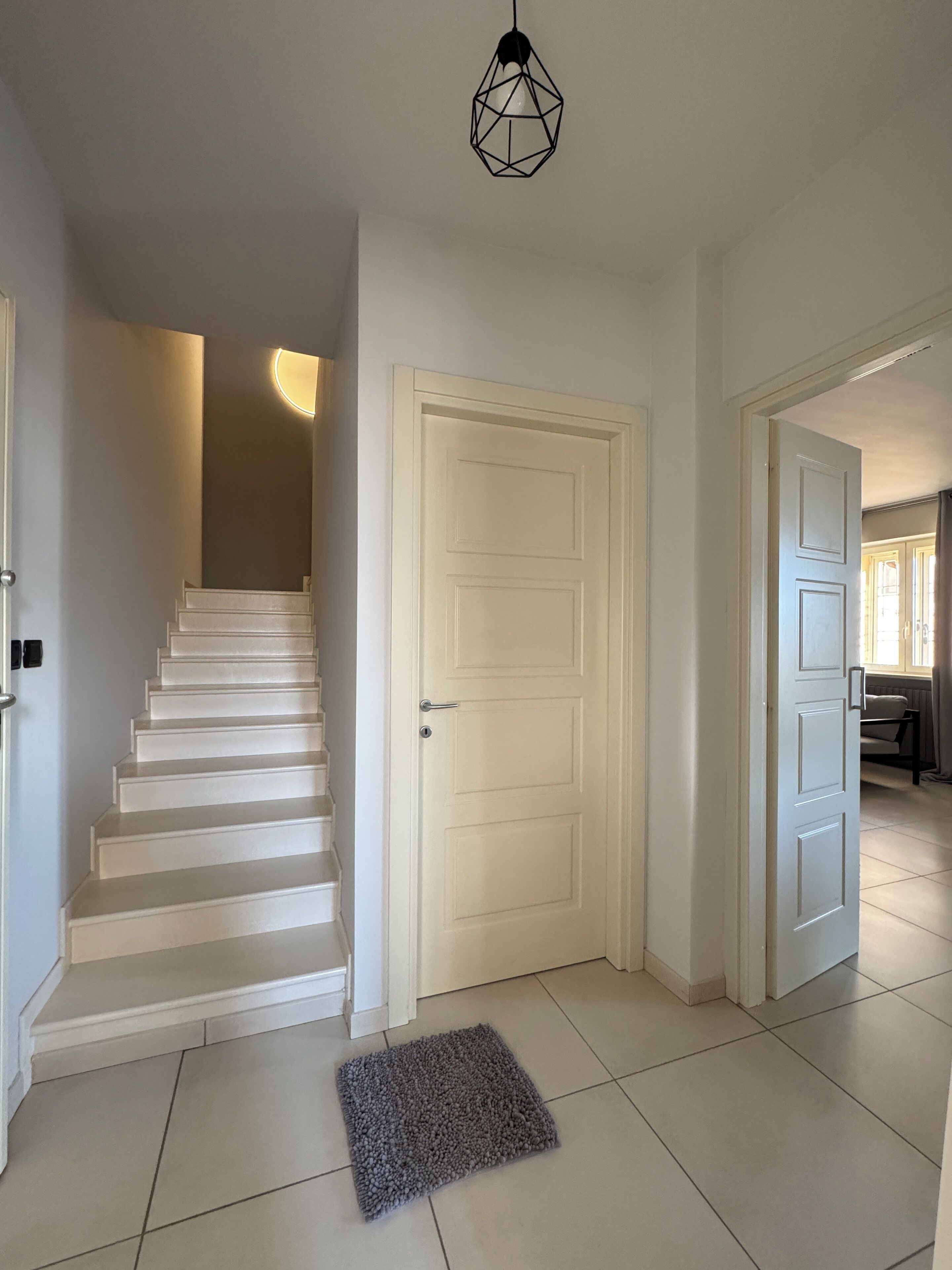 Un escalier menant à un palier avec une porte blanche, un tapis gris sur le sol et un luminaire suspendu.