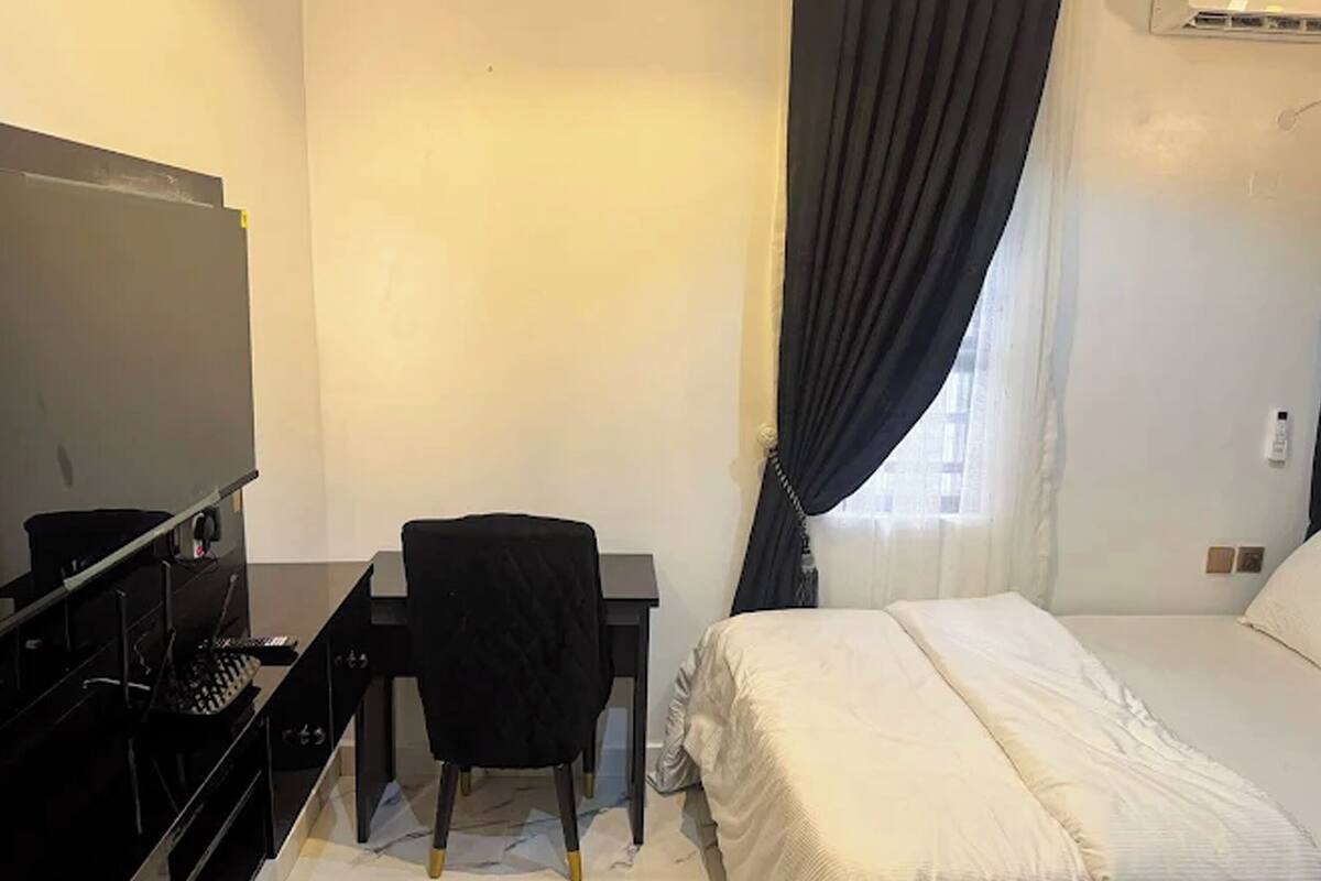 Quarto de hotel com cama, escrivaninha, cadeira, televisão e janela com cortinas.