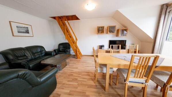 Een woonkamer met een zwarte leren bank, een houten eettafel met stoelen, een houten trap en een open haard.