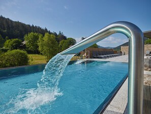 A swimming pool with a water feature, surrounded by a wooden deck and a stone wall, with a mountainous landscape in the background. - Hotel Heselbacher Hof  (Baiersbronn)