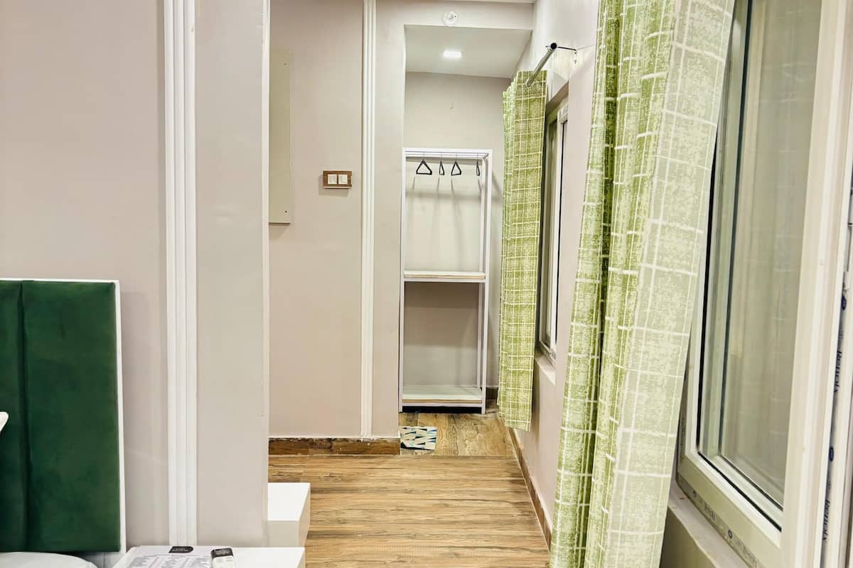 Un corridoio stretto con pavimento in legno, un comodino bianco con un telecomando, un asciugamano verde e una finestra con tende verdi.