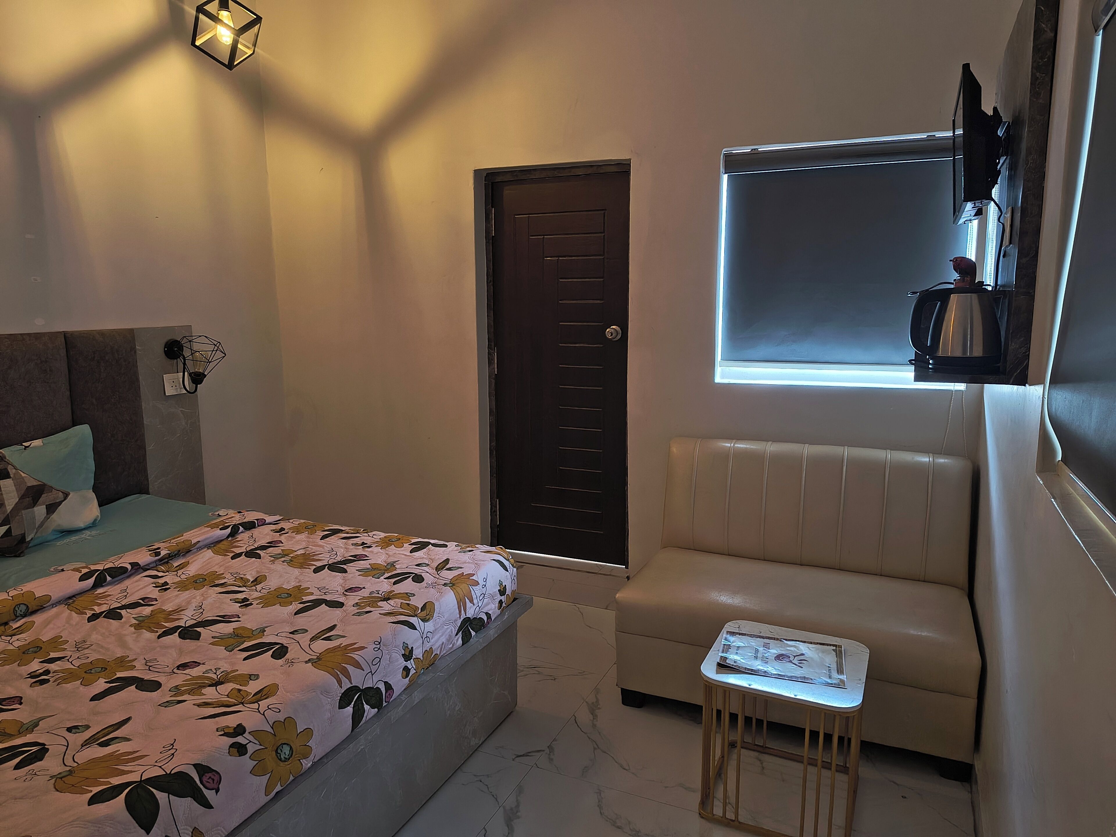 A bedroom with a bed, a chair, a small table, a window with blinds, and a door.