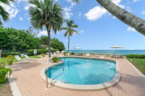 Zona de piscina con sillones de playa y sombrillas, rodeada de palmeras y vista al océano.