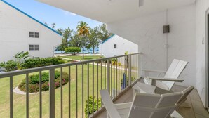 Un balcón con sillas blancas, ventilador de techo y vista a una piscina y palmeras.
