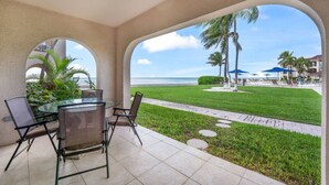 A covered patio with a glass table and chairs, a view of the beach, palm trees, and a pathway leading to the beach. - George Town Villas 117 (Seven Mile Beach)