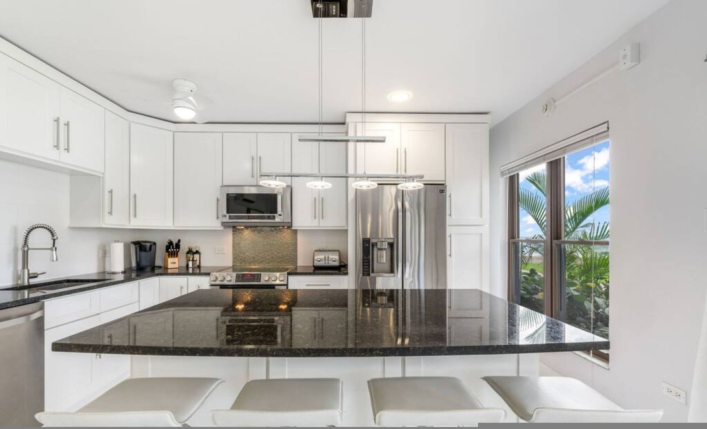 A modern kitchen with white cabinetry, a large island with seating, stainless steel appliances, and a view of palm trees through the window.
