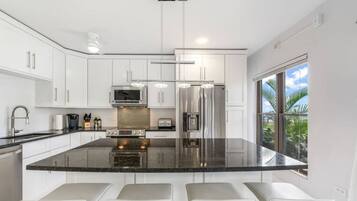 A modern kitchen with white cabinetry, a large island with seating, stainless steel appliances, and a view of palm trees through the window.