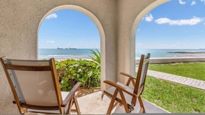Ein überdachter Sitzbereich im Freien mit zwei Holzstühlen und einem Tisch, mit Blick auf einen Strand und freie Sicht auf das Meer und ein Schiff in der Ferne.