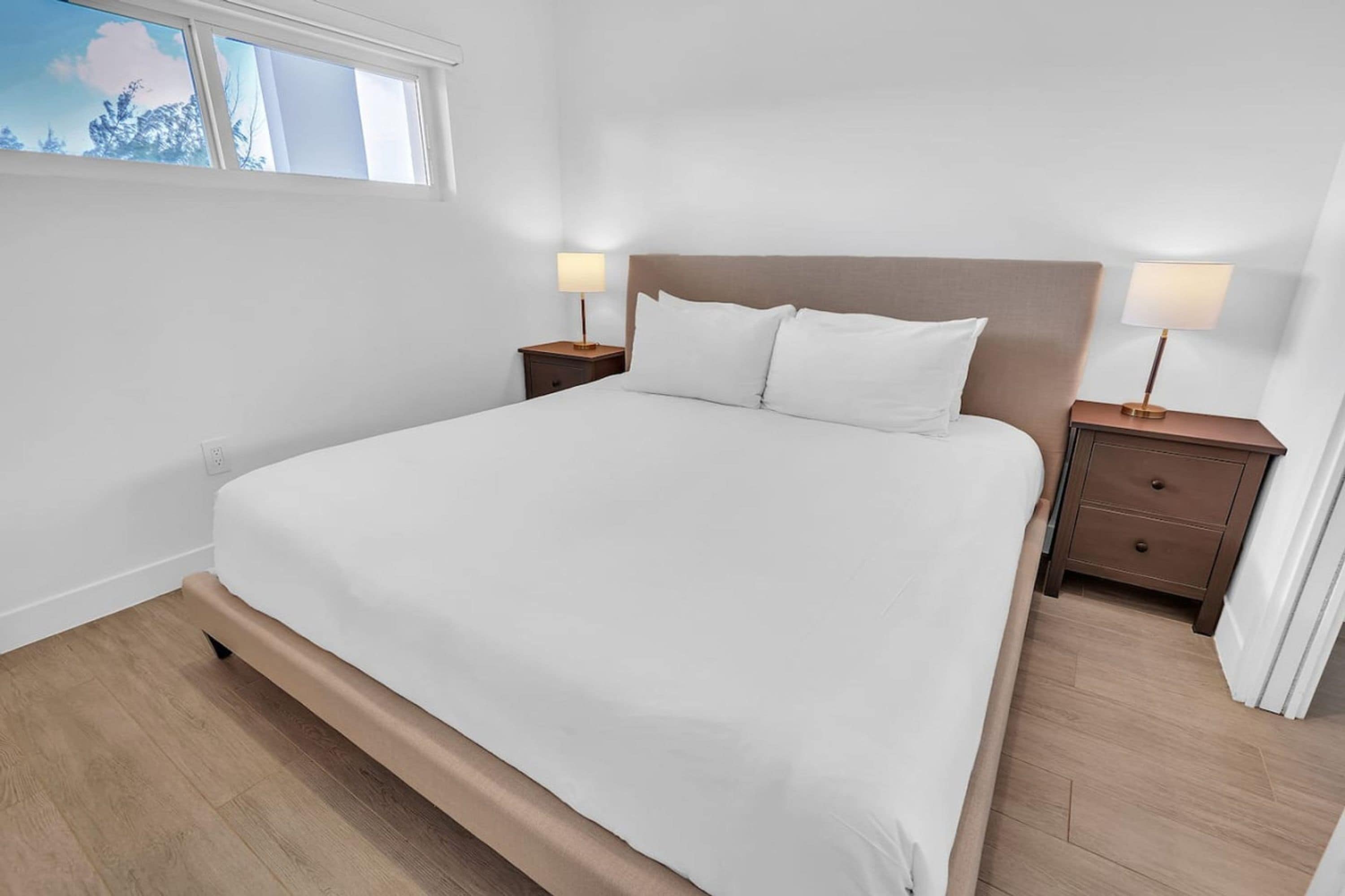A bedroom with a large bed, two bedside tables with lamps, a window with a view of trees, and a wooden floor.
