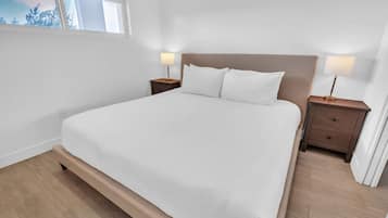 A bedroom with a large bed, two bedside tables with lamps, a window with a view of trees, and a wooden floor.