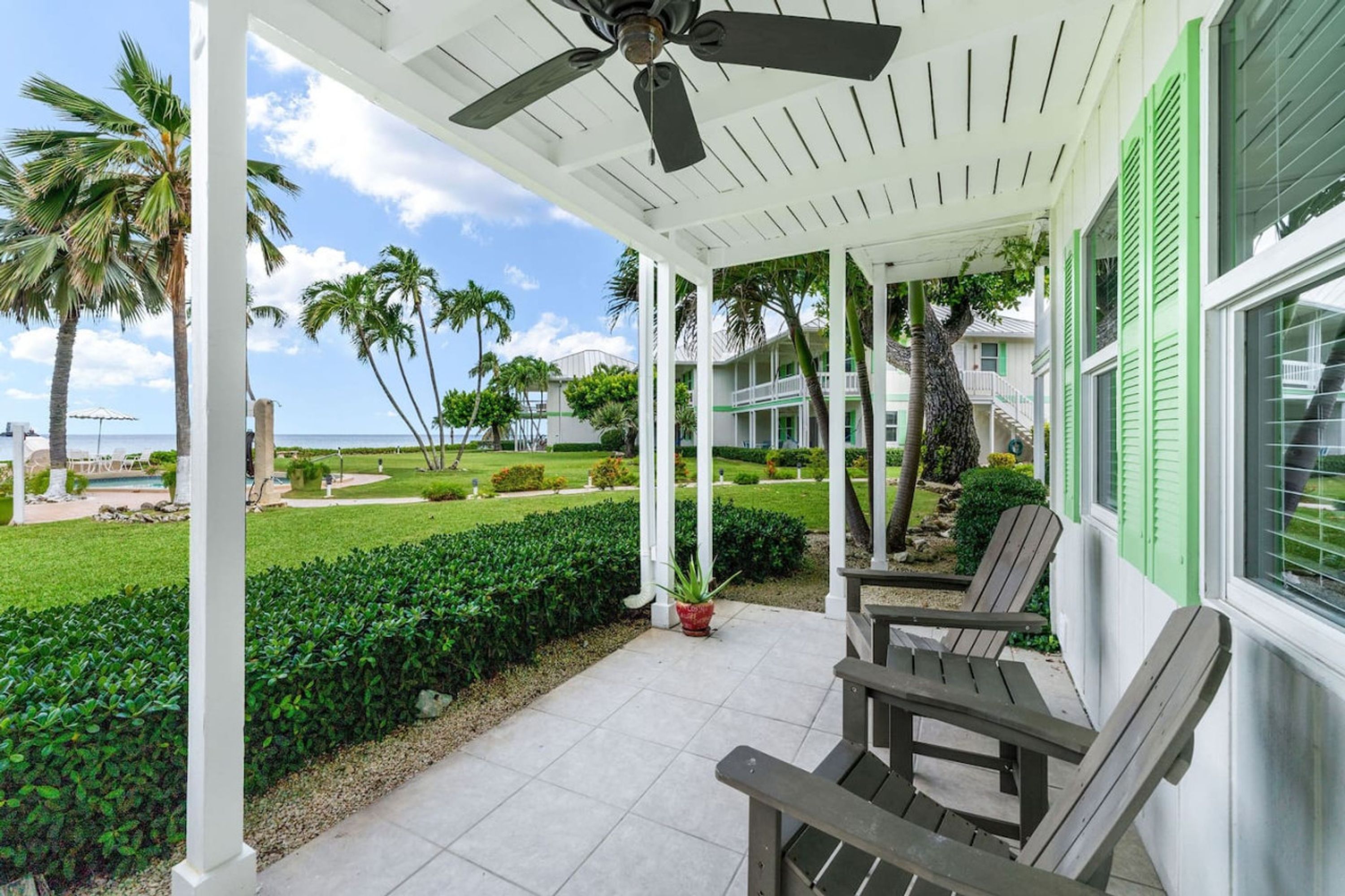 A covered outdoor area with a ceiling fan, wooden chairs, and a view of a well-maintained garden and ocean.