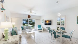 A spacious living room with a white ceiling fan, a TV mounted on the wall, and a dining area with white wicker furniture.