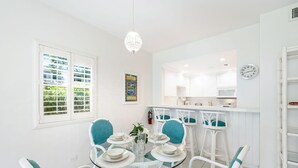 A dining area with a round glass table, white chairs, and a chandelier. There is a kitchen in the background with a clock and a window with blinds.