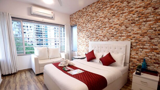 A hotel room with a brick wall, a large bed with red pillows, a white sofa, and a bedside table with a bottle.