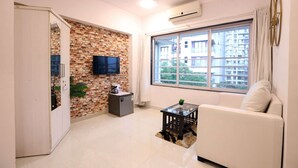 A modern living room with a white sofa, a TV mounted on a brick wall, a large window with a view of buildings, and a ceiling fan.
