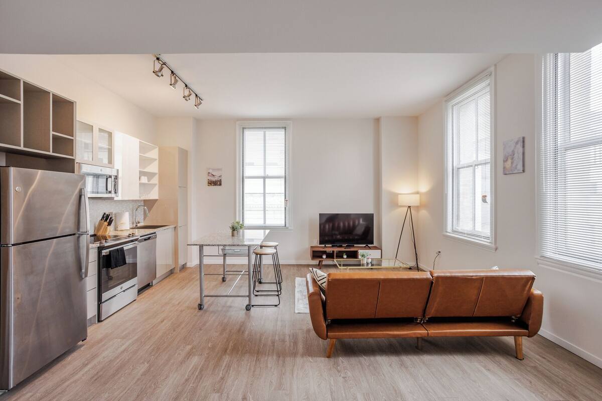 A modern kitchen with a built-in oven, microwave, and stainless steel refrigerator. There is a dining area with a wooden table and chairs, and a brown leather sofa.