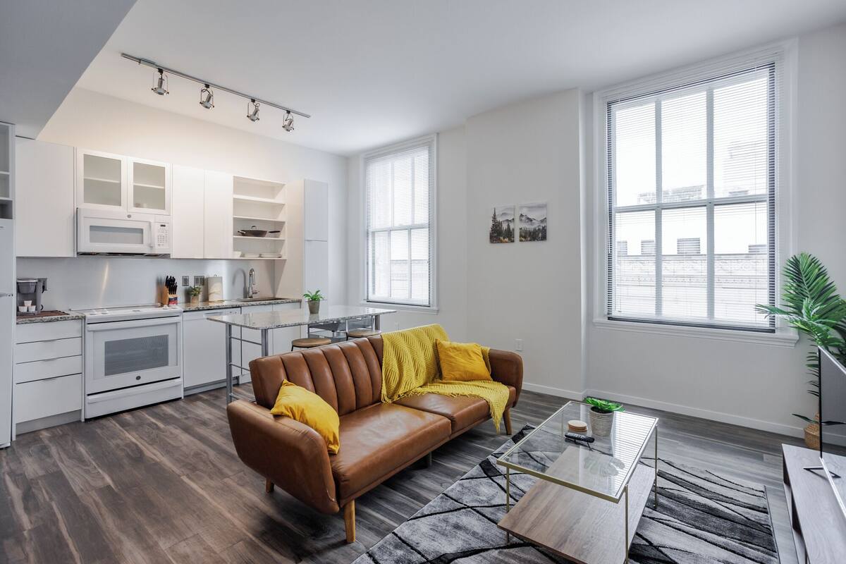 A modern apartment with a kitchen, a living area with a brown leather sofa, and large windows.