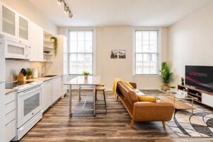 Una cocina moderna con muebles blancos y microondas integrado, un comedor con mesa y sillas, un sofá y una televisión en una sala de estar con ventanas amplias.
