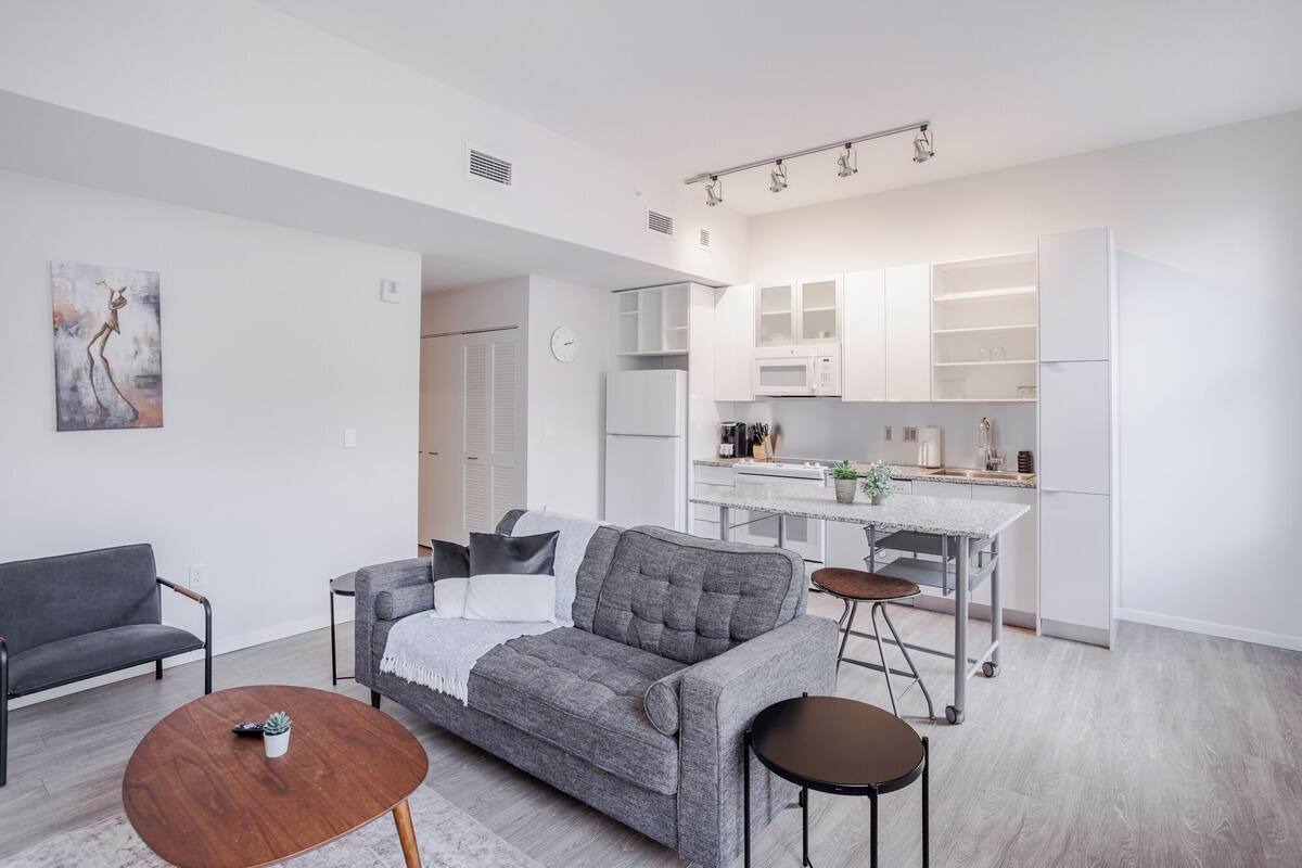 A modern living room with a grey sofa, a coffee table, and a dining area with a kitchen in the background.