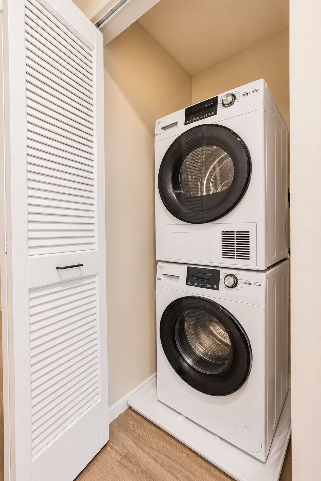 Una lavadora y secadora apiladas en un cuarto de lavado, con una puerta blanca y piso de madera.