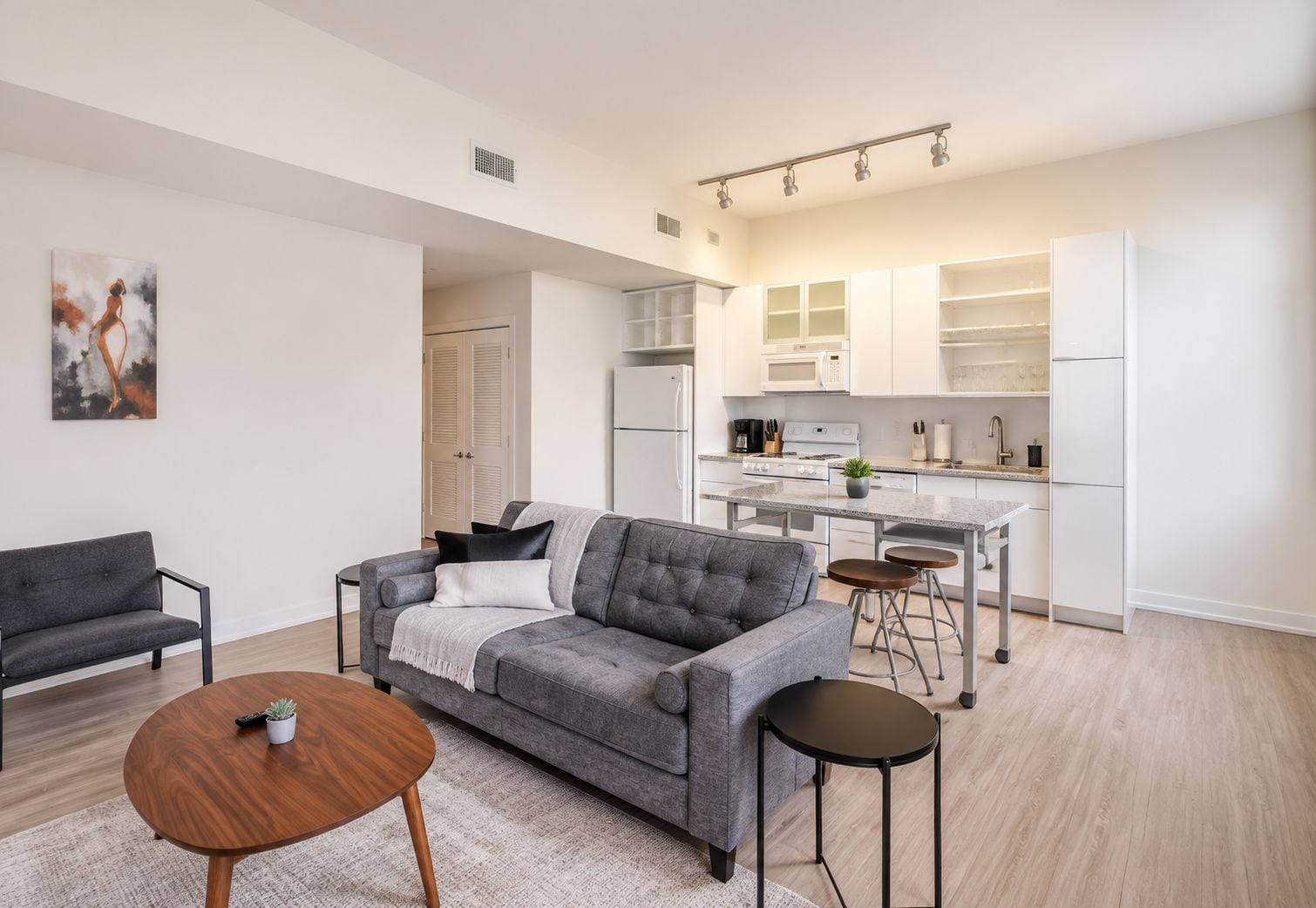 A modern apartment with a grey sofa, a wooden coffee table, a kitchen with white cabinets, and a painting on the wall.
