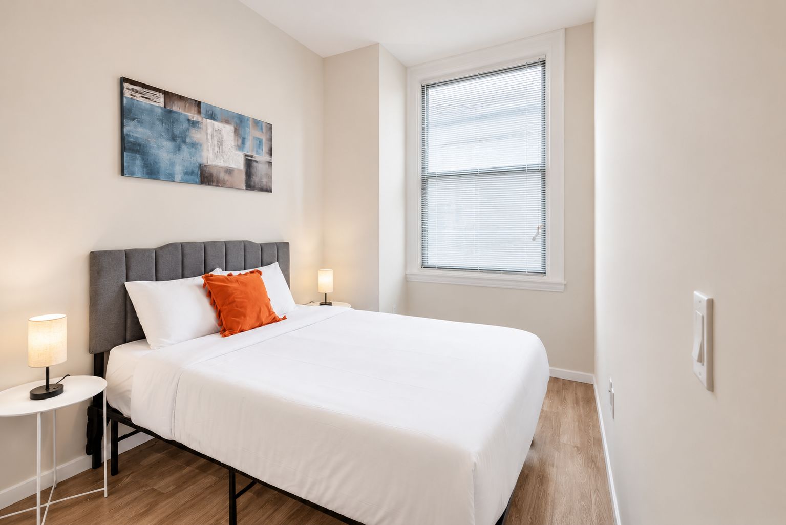 A hotel room with a bed, two bedside lamps, a window with blinds, and a painting on the wall.