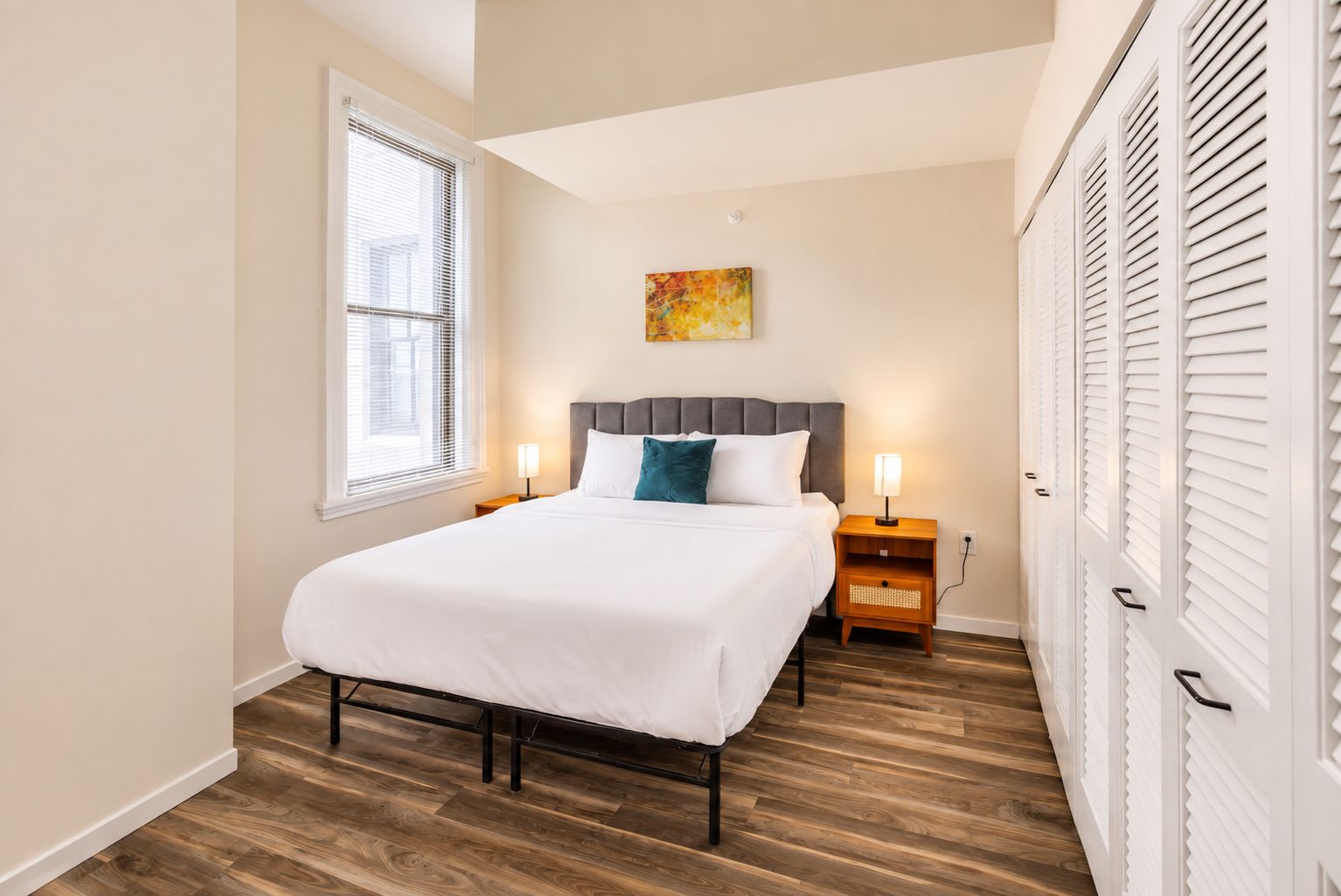 A bedroom with a bed, bedside tables, a window with blinds, and a wall-mounted artwork.