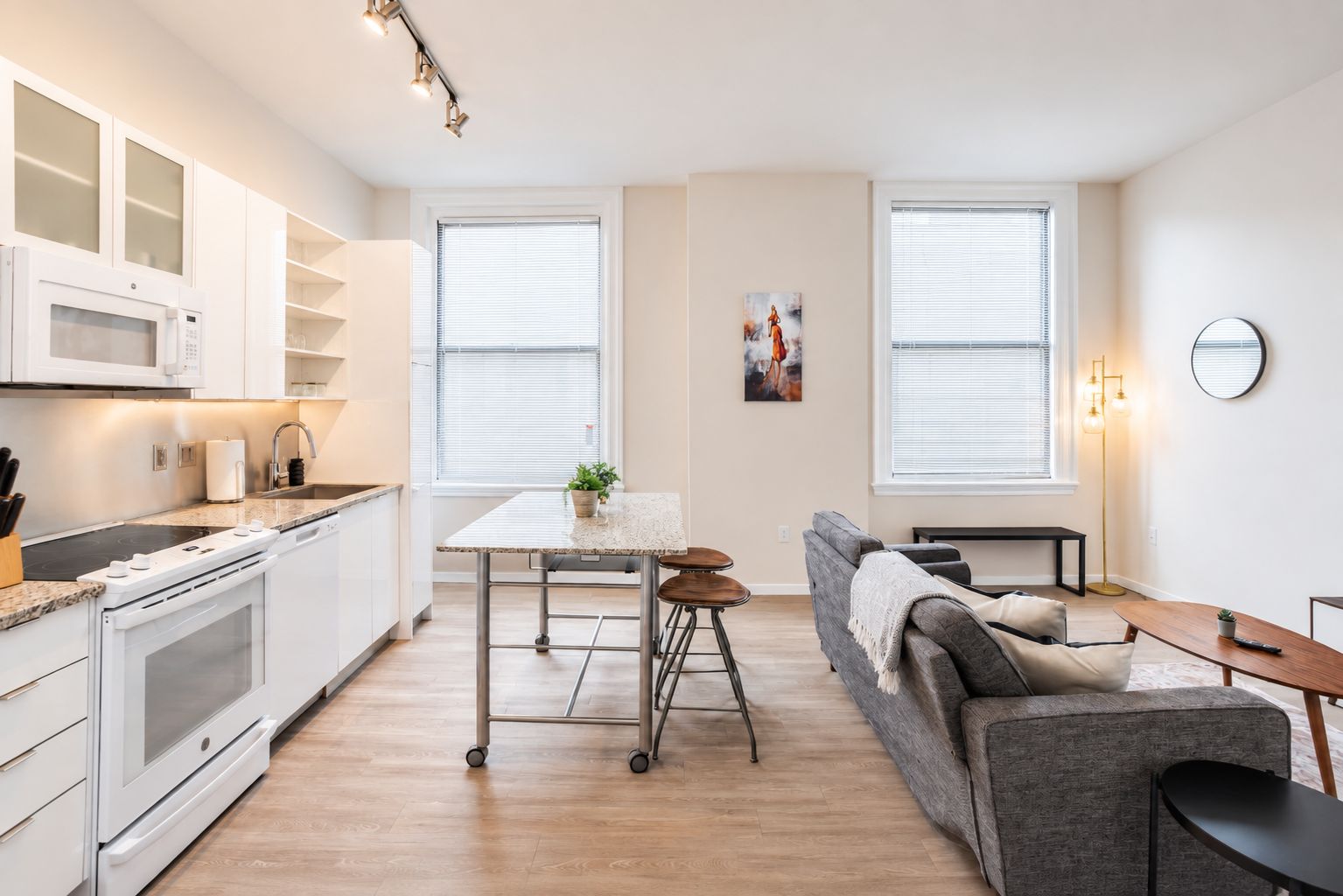 A modern kitchen with white cabinets, a microwave, and a stove. There is a dining area with a table and chairs, and a gray sofa.